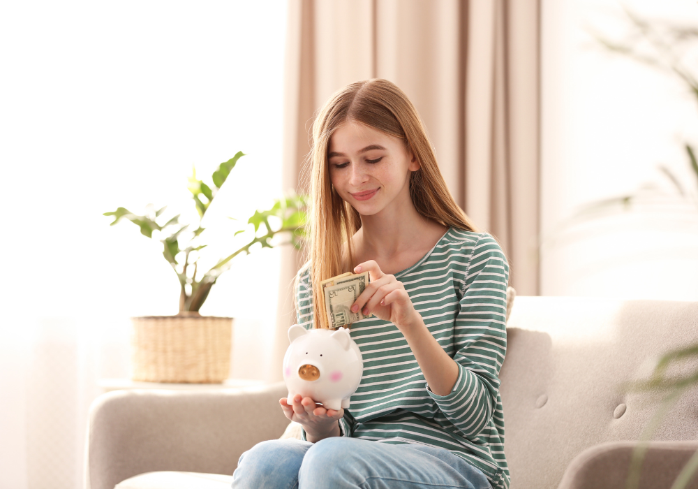 teenage girl sitting down holding money and a piggy bank